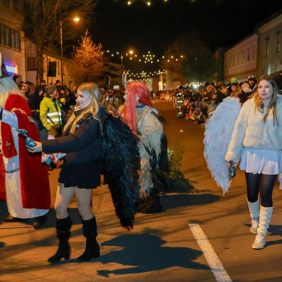 Bei Nacht zieht eine festliche Parade vorbei, mit einer Frau in einem schwarzen Outfit mit Flügeln, einem Mädchen in einem weißen Pelzmantel und einem Mann in einem roten Gewand mit einem gehörnten Kopfschmuck. Die Menge sieht gespannt zu.