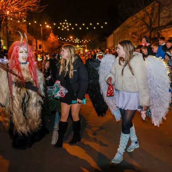Zwei Frauen gehen bei Nacht eine Straße entlang, eine in einem Engelskostüm und die andere in einem Monsterkostüm. Sie sind von anderen Leuten umgeben.