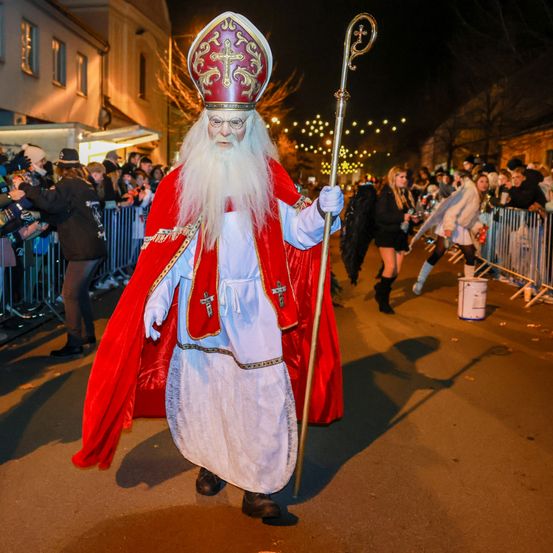Eine Person, die als Bischof verkleidet ist, zieht nachts mit einem Stab in einer Parade, während eine Menge Menschen hinter ihm jubelt.