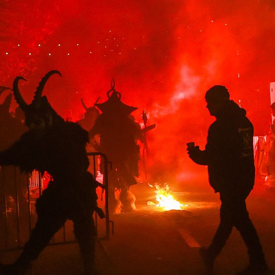 Ein Lagerfeuer brennt auf einer Straße, neben zwei Personen in Hörnerkostümen. Eine Person in einer Kapuze geht vorbei und hält einen Becher.