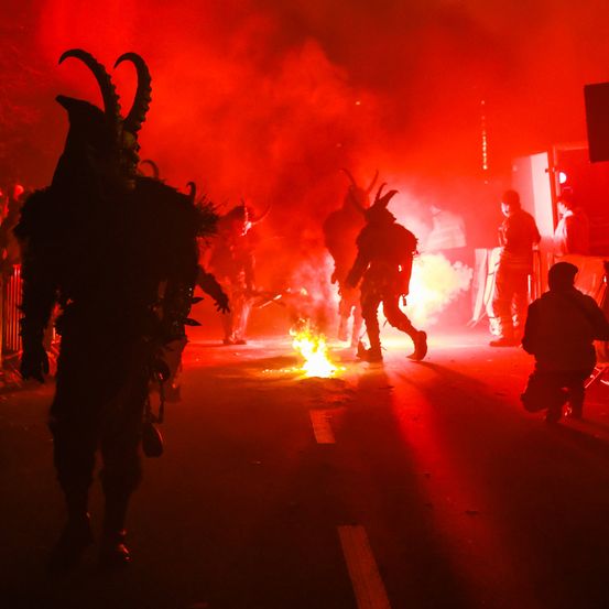 Menschen in Hörnerkostümen gehen auf einer Straße mit roten Lichtern, Feuer und Rauch im Hintergrund. Eine Person kniet und eine andere steht in der Nähe.