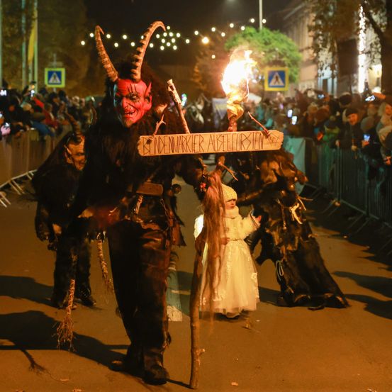 Eine Person in einem Hörnerkostüm hält ein Schild und eine Fackel, geht mit einem Mädchen in einem weißen Kleid auf einer Straße bei Nacht, umgeben von einer Menge.