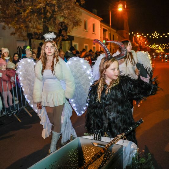 Zwei Mädchen in Kostümen gehen bei Nacht durch eine Parade. Eine ist als Engel gekleidet, die andere als Teufel. Die Straße ist mit Lichtern geschmückt, und eine Menge beobachtet sie.