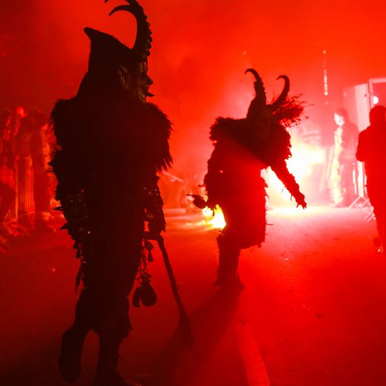 Zwei Personen in Hörnermasken und Kostümen gehen in einem rot beleuchteten Bereich, möglicherweise bei einem Festival. Ein Feuer brennt hinter ihnen.