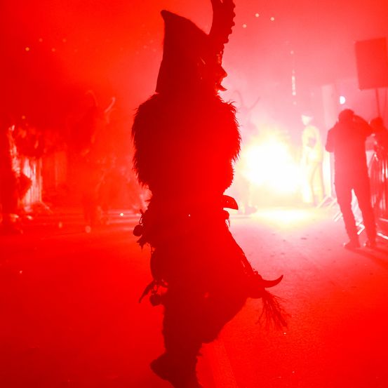 Eine Person in einem Kostüm tanzt unter rotem Licht mit einem Feuer im Hintergrund, während andere im Hintergrund stehen.