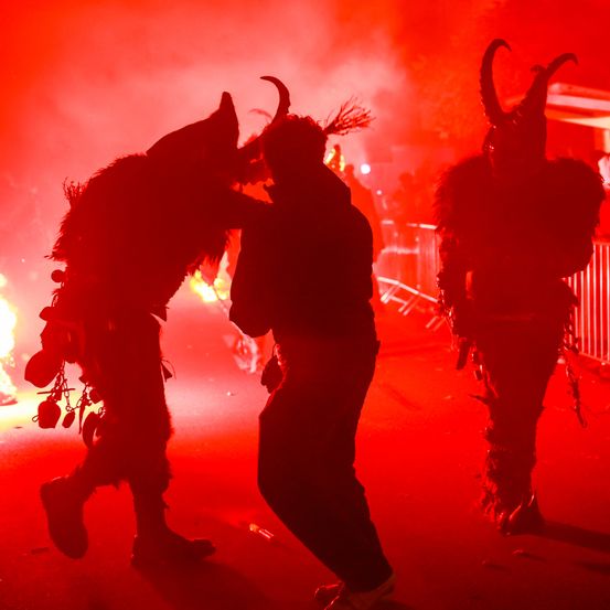 Drei Personen in Hörnerkostümen stehen auf einer roten Bühne, wahrscheinlich bei einem Festival. Sie scheinen eine Aufführung zu machen.