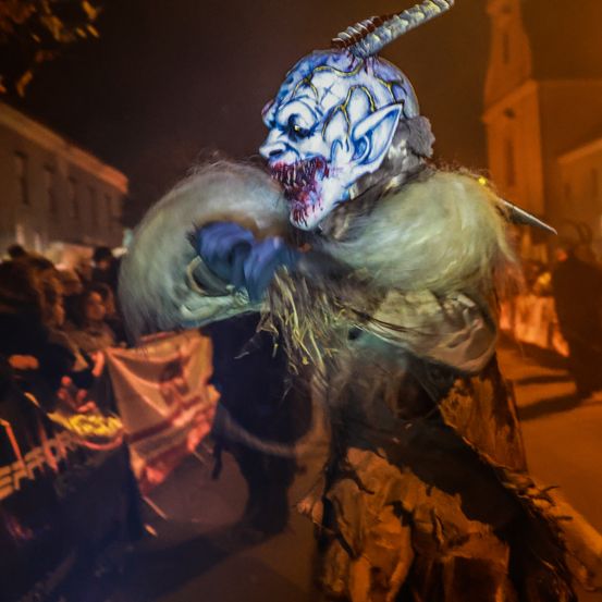 Eine Person mit einer blau-weißen Maske mit Hörnern und einem bedrohlichen Gesicht zieht bei Nacht in einem Umzug.