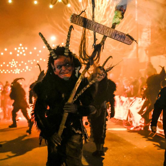 Eine maskierte Person in einem gehörnten Kostüm hält eine Fackel und ein Schild und geht in einer von Feuerschein und verstreuten Lichtern erleuchteten Straße mit anderen.