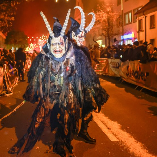 Zwei Personen in Hörnermasken und zerlumpten Kostümen gehen bei Nacht eine Straße entlang, während eine Menschenmenge hinter einer Absperrung sie beobachtet. Die Atmosphäre wird von roten Lichtern beleuchtet.