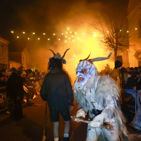Bei Nacht stehen zwei Personen in Kostümen, eine mit Hörnern und die andere mit einer Maske, vor einer Menge, mit einem Feuer und Feuerwerk im Hintergrund.