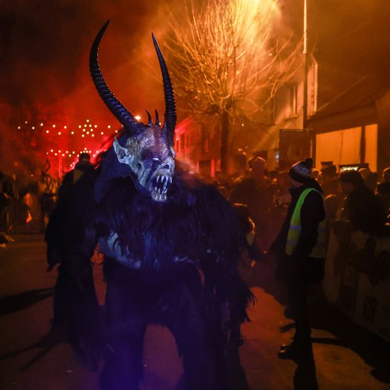 Eine Person in einem Monsterkostüm mit Hörnern läuft nachts auf einer Straße, umgeben von einer Menge und roten Lichtern.
