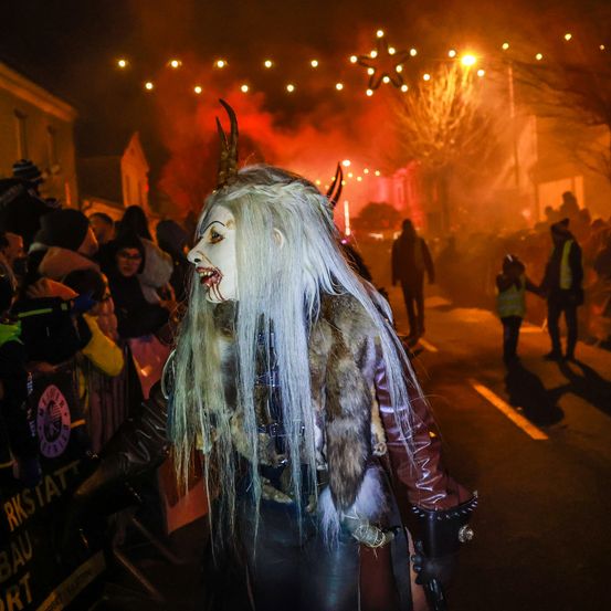 Eine Person in einer weißen Perücke und einem gehörnten Masken geht während eines Festivals mit Feuerwerk und Zuschauern eine Straße entlang.