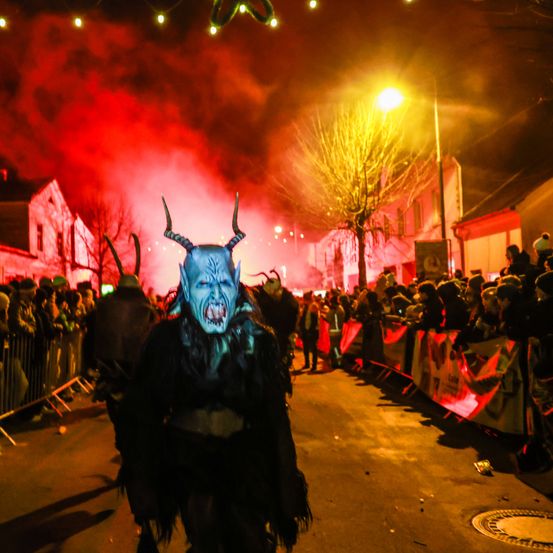 Eine Person in einer blauen Dämonenmaske mit Hörnern geht bei Nacht die Straße entlang, umgeben von einer Menge mit rotem Rauch und Lichtern im Hintergrund.