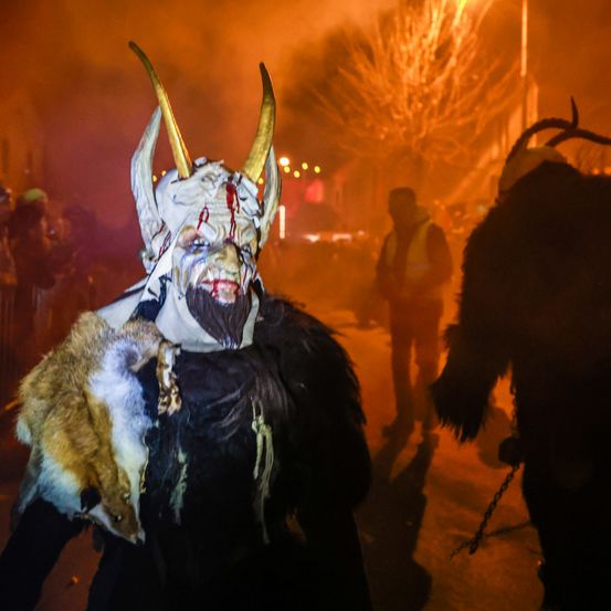 Eine Person mit Hornmaske geht auf einer schwach beleuchteten Straße, eine Kette im Vordergrund. Eine weitere Person ist im Hintergrund teilweise sichtbar.