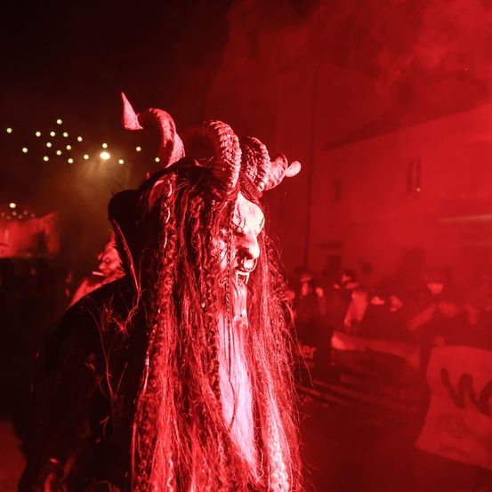 Eine Person, die als Teufel mit Hörnern und Dreadlocks verkleidet ist, steht in einer Menschenmenge. Rote Lichter beleuchten die Szene.