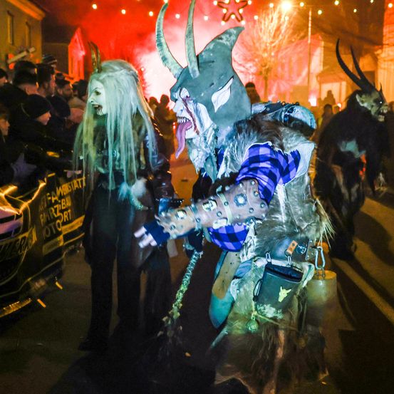 Ein nächtlicher Straßenumzug mit kostümierten Teilnehmern in Hörnerhelmen und Ketten, inmitten einer Menge und festlicher Lichter.