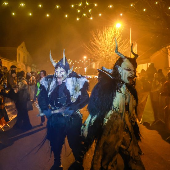 Zwei Menschen in Hörnermasken gehen in einer Nachtstraße, hinter ihnen eine Menge, beleuchtet von warmen orangefarbenen Lichtern.