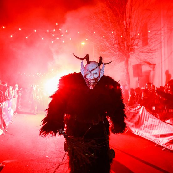 Eine Person in einem Dämonenkostüm mit Hörnern und einer Maske geht einen roten Teppich hinunter, während eine Menge auf beiden Seiten steht. Im Hintergrund sind rote Lichter und Rauch zu sehen.