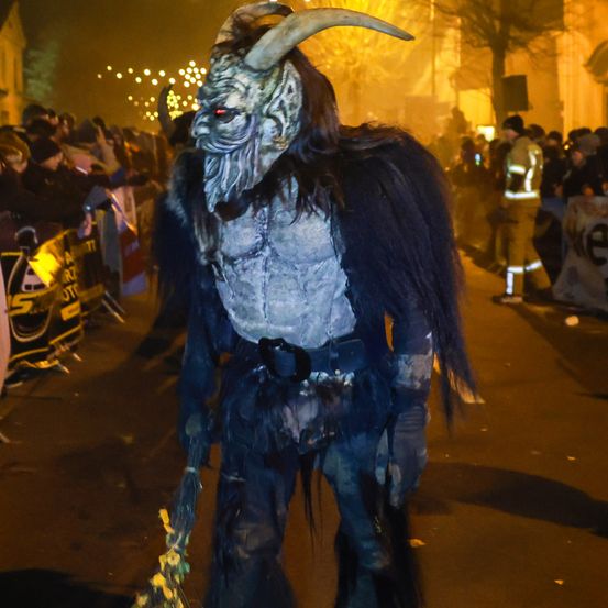 Eine Person in einem gruseligen Kostüm mit Hörnern und einer Maske geht bei Nacht auf der Straße. Es gibt Zuschauer und einen Feuerwehrmann, der hinter einer Barriere steht.