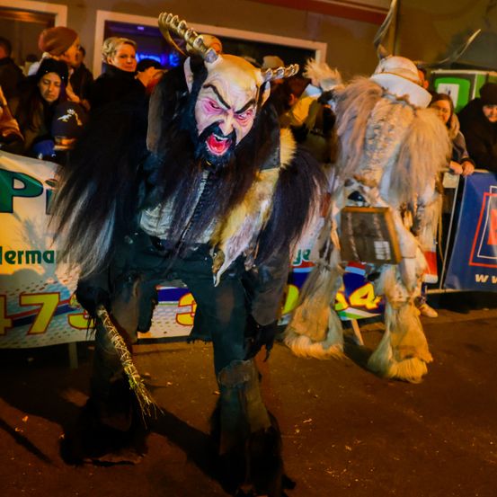 Eine Person in einem Dämonenkostüm steht vor einer Menge. Die Menge beobachtet die Person. Hinter der Menge befindet sich ein Banner.