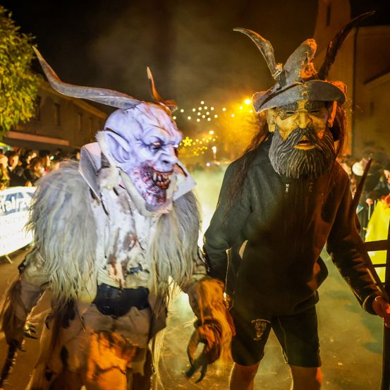 Zwei Personen in aufwendigen Kostümen gehen bei einer Nachtparade. Einer trägt eine Hörnermaske, der andere eine Bartmaske. Sie werden von einer Menschenmenge begleitet.