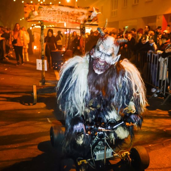 Eine Person in einem grotesken Kostüm mit Maske und Hörnern fährt nachts in einem Umzug auf einem Dreirad. Die Menge steht hinter einem Metallzaun. Ein Schild mit einer fremden Sprache ist über ihnen.
