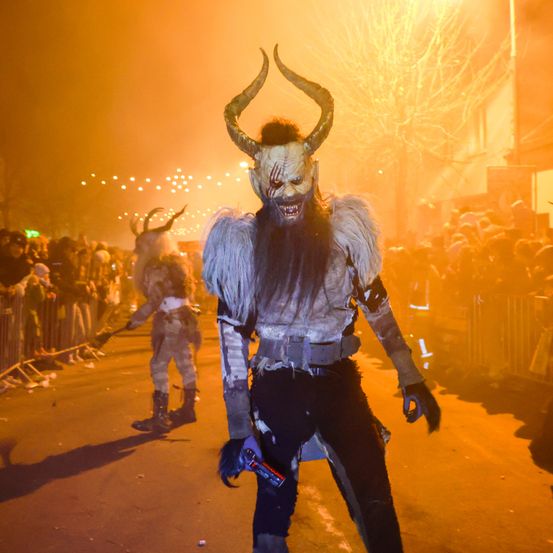 Eine Person in einem Dämonenkostüm mit Hörnern steht auf einer Straße während eines Festivals, während eine Menge zuschaut.