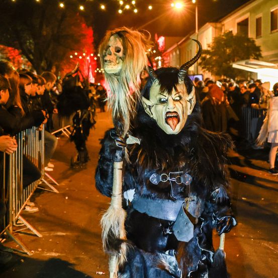 Eine Person, die als Teufel mit Hörnern und Maske verkleidet ist, steht bei Nacht vor einer Menschenmenge. Die Person hält eine Puppe mit einem schädelartigen Gesicht.