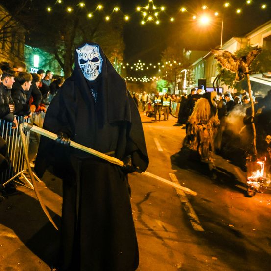 Eine Person mit Totenkopfmaske und schwarzem Gewand, die eine Sense hält, steht in einer Menschenmenge auf einer Straße bei Nacht.