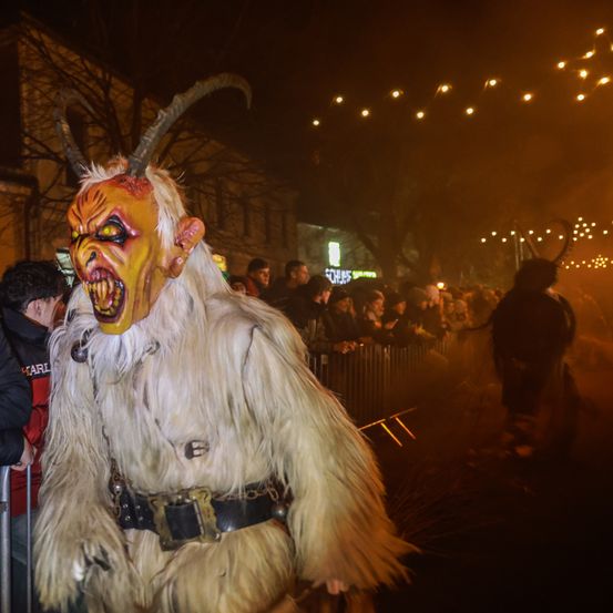 Eine Person in einem hörnertragenden Dämonenkostüm, umgeben von einer Menge, während eines nächtlichen Festivals. Der Kostüm ist weiß mit einer gelben Maske, und der Hintergrund ist dunkel mit beleuchteten Lichtern.