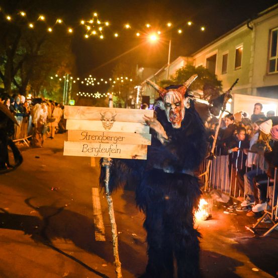 Eine Person in einem Kostüm mit Hörnern steht auf der Straße und hält ein Schild mit der Aufschrift 'Strenoberoer Bergteufel'. Dahinter befindet sich eine Menschenmenge hinter einem Zaun, mit Straßenlaternen und Gebäuden im Hintergrund.