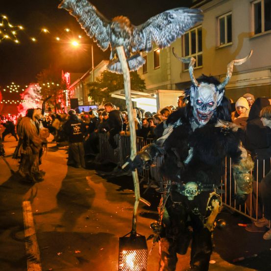 Eine Person in einem Dämonenkostüm zieht bei Nacht mit einer Laterne und einem Stab mit einem Adler auf der Spitze in einer Parade, während eine Menge zuschaut.