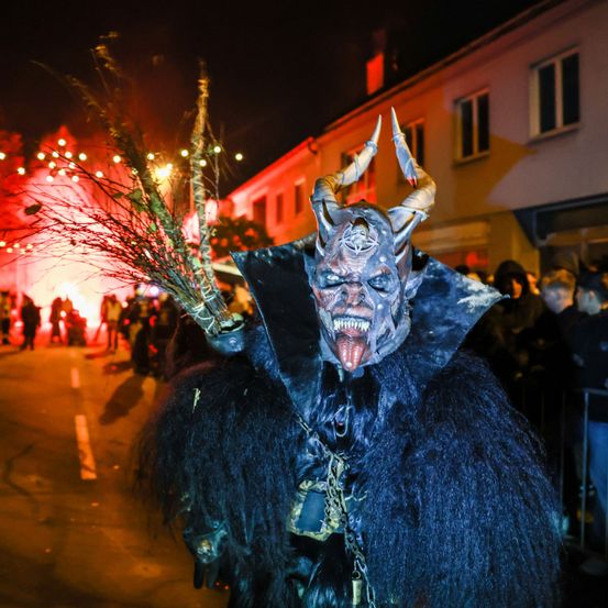 Eine Person in einem Dämonenkostüm steht bei Nacht auf einer Straße, umgeben von Menschen, mit Gebäuden und Feuer im Hintergrund.