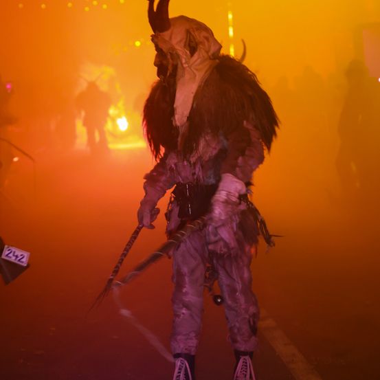 Eine Person in einer gehörnten Maske und Kostüm steht in einem orange beleuchteten Bereich, möglicherweise bei einem Festival. Der Hintergrund ist unscharf, und eine Nummer 242 ist sichtbar.