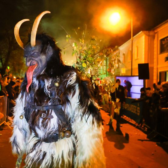 Eine Person in einem pelzigen Kostüm mit Hörnern und einer roten Zunge läuft bei Nacht in einer Parade. Im Hintergrund befindet sich ein Gebäude mit einer großen Leinwand.
