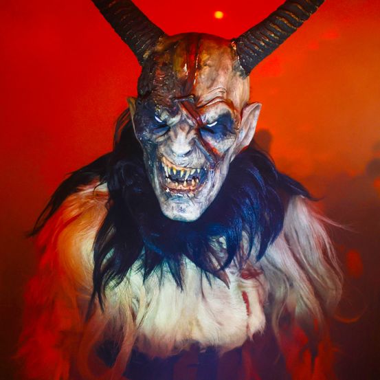Eine Dämonenmaske mit großen Hörnern und einem bedrohlichen Ausdruck, in Weiß und Blau bemalt. Die Maske hat einen roten Hintergrund.