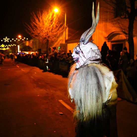 Eine Person in einem gruseligen Monsterkostüm geht bei Nacht auf der Straße. Viele Menschen schauen von hinter einer Barriere zu.