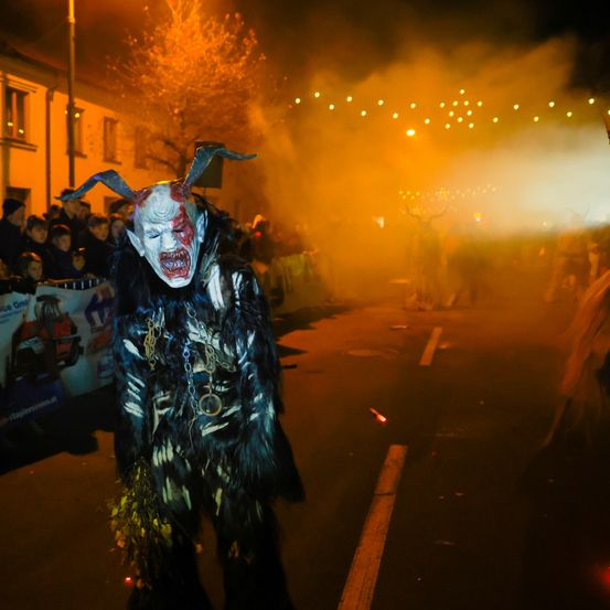 Eine Person in einer Hörnermaske und Kostüm geht bei Nacht auf einer Straße, mit Rauch und Lichtern im Hintergrund, während ein Publikum zuschaut.