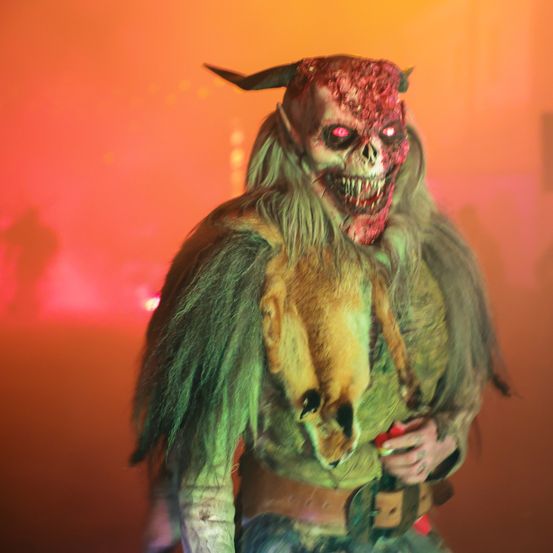 Eine dämonische Figur mit Hörnern und einer blutigen Maske steht vor einem verschwommenen orangefarbenen Hintergrund. Die Figur trägt ein zerlumptes, grünes Kostüm mit einem toten Tier auf der Brust.