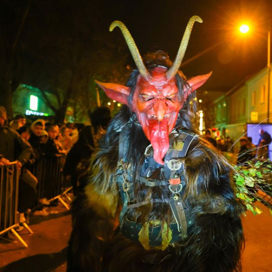 Eine Person, die als Dämon mit Hörnern und einer roten Maske verkleidet ist, steht nachts auf der Straße, während eine Menschenmenge hinter einem Zaun zuschaut.