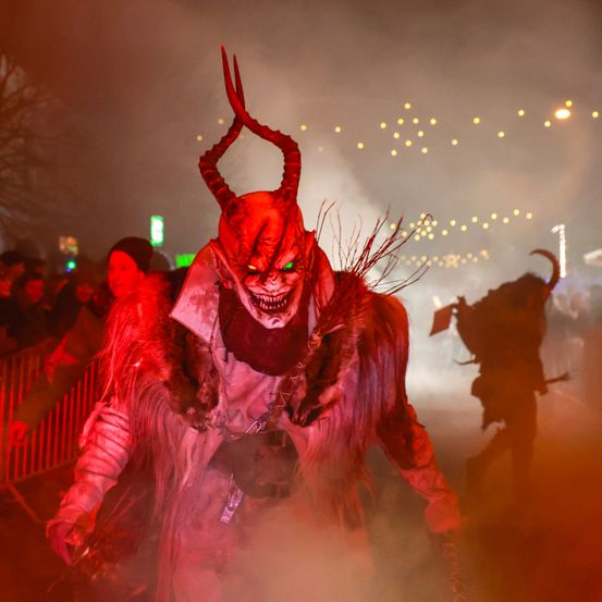Eine Person in einem teuflischen Kostüm mit Hörnern und leuchtenden Augen geht bei Nacht durch eine Menschenmenge. Der Hintergrund ist dunkel mit roter Beleuchtung.