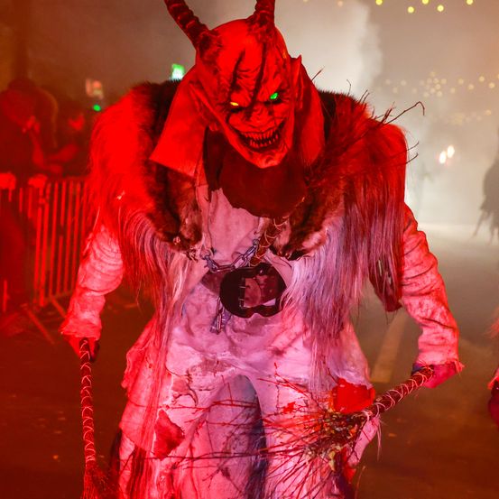 Eine Person in einer Dämonen-Verkleidung mit roter Maske, leuchtend grünen Augen und Hörnern steht in einem schwach beleuchteten Bereich. Die Person hält einen blutbefleckten Besen in beiden Händen.