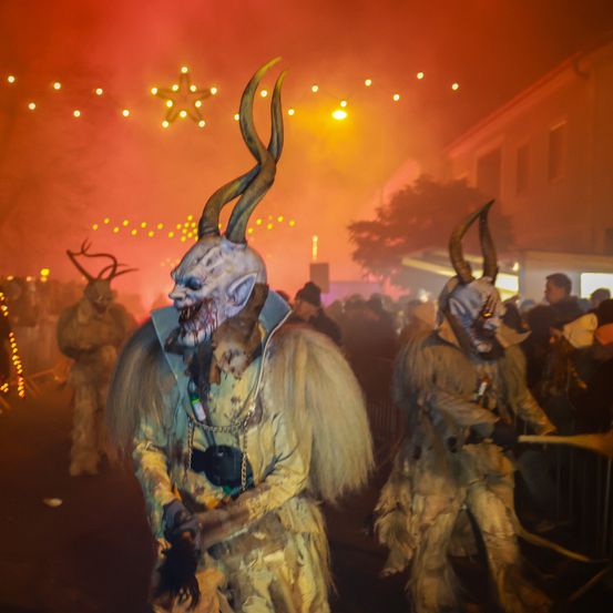 Eine Menschenmenge in hörnertragenden Teufelskostümen zieht in einer Parade, umgeben von Feuer und Rauch.