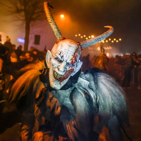 Eine Person in einer Hörnermaske und Fellkostüm steht bei Nacht in einer Menschenmenge. Die Szene wird von orangefarbenen Lichtern beleuchtet, mit entfernten Menschen und Strukturen im Hintergrund.