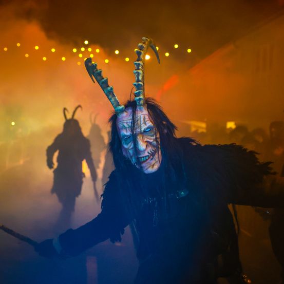 Eine Person mit einer Hörnermaske steht in einem schwach beleuchteten Raum und hält ein Schwert. Hinter ihr ist eine weitere Figur teilweise sichtbar im Hintergrund.