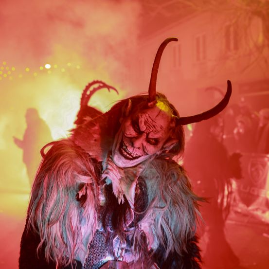 Eine Person in Teufelskostüm mit Hörnern und einer furchterregenden Maske, die in einem rot beleuchteten Bereich steht, wahrscheinlich für eine Halloween-Veranstaltung.