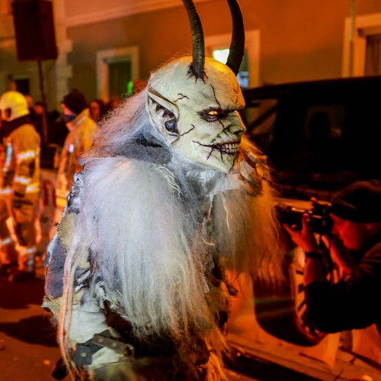 Eine Person in einem weißen und grauen Kostüm mit Hörnern, die bei Nacht in einem Umzug geht. Ein Fotograf hält die Szene mit einer Kamera fest. Dahinter stehen Menschen in Schutzanzügen und beobachten.