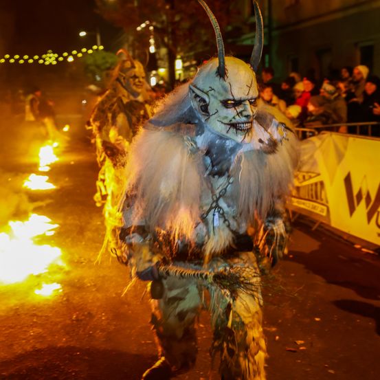 Zwei Personen in Monsterkostümen gehen bei Nacht auf einer Straße, umgeben von Zuschauern und brennenden Feuern.