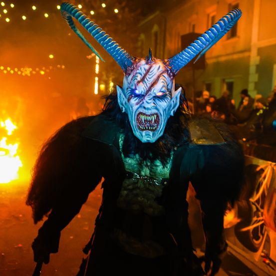 Eine Person in Teufelsverkleidung mit Hörnern und blauen Lichtaugen steht vor einem Feuer, wahrscheinlich bei einer Halloween-Veranstaltung.
