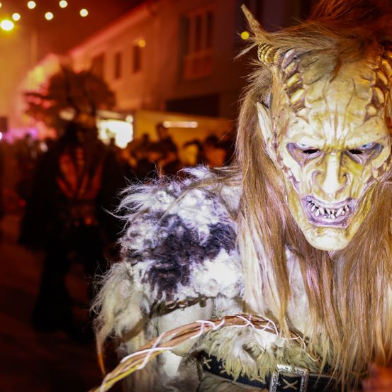 Eine Person mit einer hörnermaskierten Monstermaske läuft nachts auf einer Straße. Im Hintergrund gehen mehrere Leute. Es gibt Gebäude mit Lichtern.